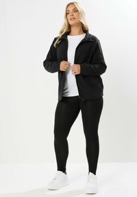 Svart fleece zipjacka över en vit skjorta, kombinerad med svarta tights och vita sneakers. Slät textur, avslappnad passform och detaljerad krage.