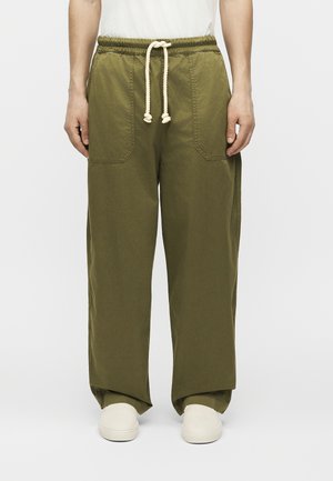 Άτομο που φορά φαρδιά ελιάς πράσινα παντελόνια με μεγάλες μπροστινές τσέπες και λευκό κορδόνι, σε συνδυασμό με λευκά παπούτσια χωρίς κορδόνια.