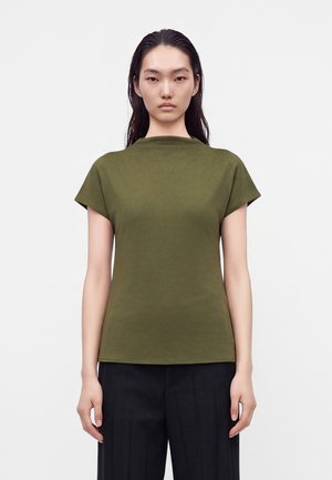 Une femme porte un haut vert olive à manches courtes et à col montant, ainsi qu’un pantalon noir, sur un fond uni et clair.