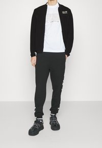 Giacca nera con zip e colletto alto, t-shirt bianca con logo, e pantaloni jogger neri con strisce laterali bianche. Sneakers nere con lacci.