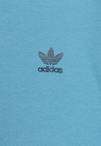 Ljusblått stickat tyg med en texturerad yta som har en mörkgrå Adidas-logotyp med trebladig design och horisontella ränder under.