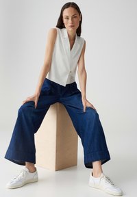 Blazer bianco senza maniche con scollatura a V e dettaglio con un singolo bottone, abbinato a pantaloni in denim blu scuro a gamba larga e sneakers bianche.