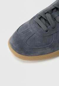 Pilkas zomšinis sportinis batelis su apvalia nosimi, audinio raišteliais ir tekstūruotu guminiu padu. Ant viršaus matomos siuvimo detalės.