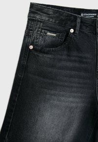 Jean en denim noir en gros plan montrant une poche avant, une petite étiquette métallique de marque, une boucle de ceinture et des rivets en cuivre sur la texture du tissu.
