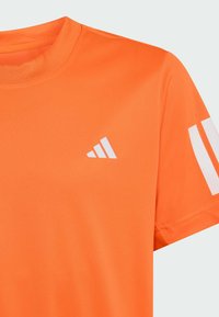 Oranžové sportovní tričko s krátkými rukávy, vyrobené z hladké tkaniny. Má bílé logo na hrudi a dvě bílé pruhy na rukávu.