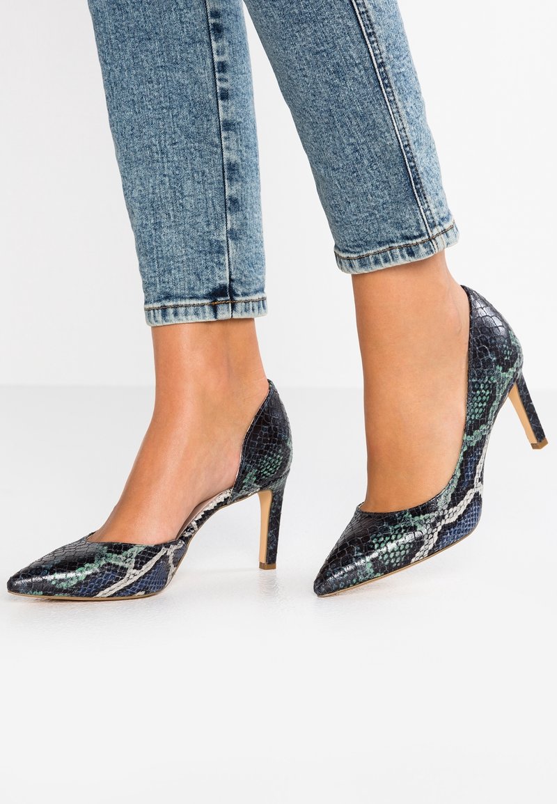 Pieds chaussés d’escarpins à bout pointu avec un motif cuir de serpent noir et vert, associés à un jean skinny bleu clair avec revers.