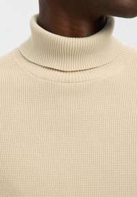 Close-up van een donkerhuidige persoon met een beige geribde coltrui tegen een effen witte achtergrond.