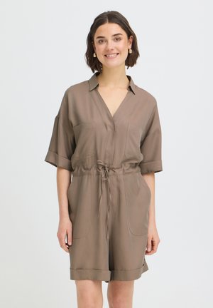 Lühikeste pruunide juustega naine kannab taupe värvi lühikeste varrukatega kraega kombinesooni, millel on nöör vöökohal, seisab ja naeratab ühevärvilise tausta ees.