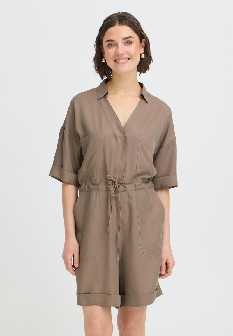 Femme aux cheveux courts bruns portant une combinaison taupe à manches courtes avec col et cordon à la taille, debout et souriante sur un fond uni.