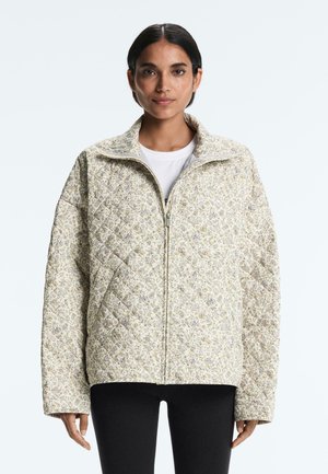 Frau trägt eine hellfarbene, gesteppte Jacke mit kleinem Blumenmuster, die hochgeschlossen ist, kombiniert mit schwarzer Hose und einem weißen Hemd darunter.