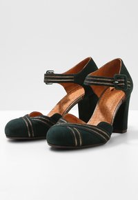 Chaussures à talons hauts en daim vert foncé avec des rayures en accent doré, bout rond et bride réglable à la cheville. Intérieur avec texture semblable à du bois.