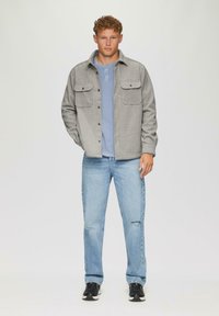 Graue Button-Down-Jacke mit zwei Brusttaschen, über einem hellblauen Henley-Shirt, kombiniert mit hellblauen Jeans und schwarzen Sneakers.