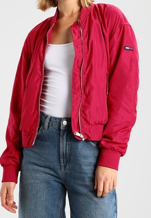Vrouw draagt een felroze bomberjack over een wit tanktop met blauwe high-waisted jeans, staand tegen een effen achtergrond.