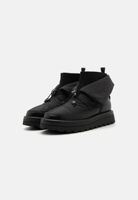 Botine negre până la gleznă, cu un superior din material matlasat, talpă din cauciuc și două corzi ajustabile pentru închidere. Include manșete elastice ribbate, înalte.
