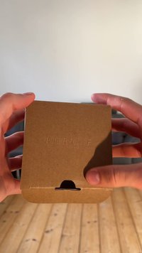 Cutie de carton maro ținută în mâini, având text în relief "PIER ONE," o deschidere fante la bază și un design simplu pliat.