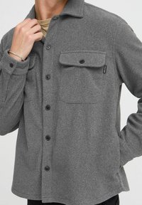 Gray button-up shirt made of textured fabric, featuring a collar, two front pockets, and black buttons. Suitable for layering.