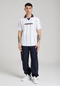 Polo bianca con righe verticali nere e colletto con accenti rossi e blu, abbinata a pantaloni scuri e sneakers bianche.