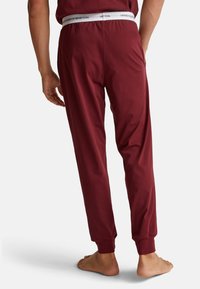 Muž v burgundských joggingových kalhotách s elastickým bílým pasem, na kterém je nápis "United Colors of Benetton", stojící bos a otočený zády.