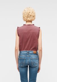 Personne aux cheveux blonds courts et bouclés, portant un haut bordeaux sans manches et un jean bleu en denim, debout de dos devant un fond uni.