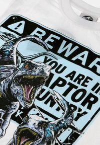 Bílé tričko s grafikou dvou dravců s detailními šupinami, ostrými zuby a zářivě oranžovými očima na světle modrém pozadí.