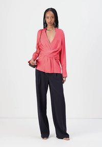 Blusa avvolgente corallo con scollatura a V profonda e maniche lunghe abbinata a pantaloni larghi a vita alta blu navy, indossata con una borsa piccola.