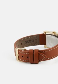 Bracelet de montre en cuir marron avec des coutures crème, boucle en métal doré et boîtier en or carré. Présente un verre transparent sur le cadran.