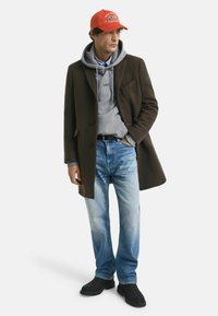 Manteau marron, hoodie gris avec logo, jeans bleus, bottes noires et casquette de baseball rouge avec logo. Le modèle se tient devant un fond blanc.