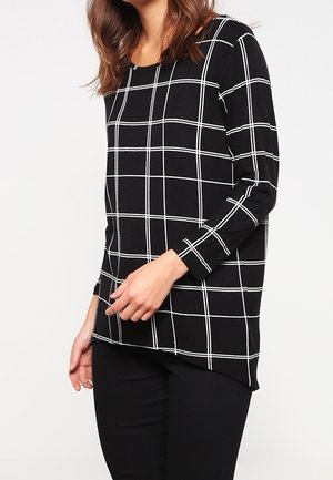 Vrouw die een zwart top met lange mouwen met wit rasterpatroon en zwarte broek draagt, staand tegen een effen achtergrond.