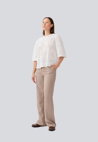 Blouse blanche en broderie anglaise avec des manches bouffantes courtes, associée à un pantalon beige. La blouse présente des motifs découpés ; le pantalon a une coupe droite avec une texture lisse.