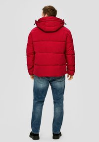 Rote Pufferjacke mit Kapuze, gepunktetem Design und elastischen Bündchen, kombiniert mit blauen Jeans und schwarzem Schuhwerk.
