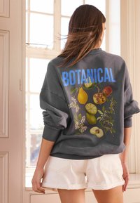 Felpa grigia con la scritta "BOTANICAL" in blu, caratterizzata da illustrazioni di frutta colorata sul retro. Indossata con pantaloni corti bianchi arrotolati.