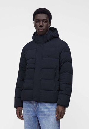 Jeune homme portant une veste matelassée noire avec capuche et un jean bleu clair, debout devant un fond clair uni.