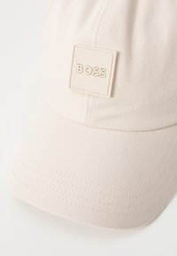 Hele roosa baseballi müts, mis on valmistatud tekstureeritud kangast ja millel on ruudukujuline logo silti, mille keskel on embosseeritud "BOSS". Kumer ääreosa koos õmblusdetailidega.