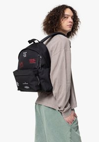 Mochila negra con bolsillo frontal, texto rojo y gráfico de corona, material duradero, correas ajustables y acabado texturizado.