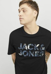 Mustapaita, jossa on lyhyet hihat ja joka esittelee tekstiä "JACK & JONES" värikkäällä kukkakuviolla etupuolella. Pehmeää kangasta ja pyöreä kaula-aukko.