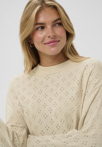 Pull en maille de couleur crème avec une coupe ample, présentant un motif texturé de petits trous sur l'ensemble et un col rond en côte.