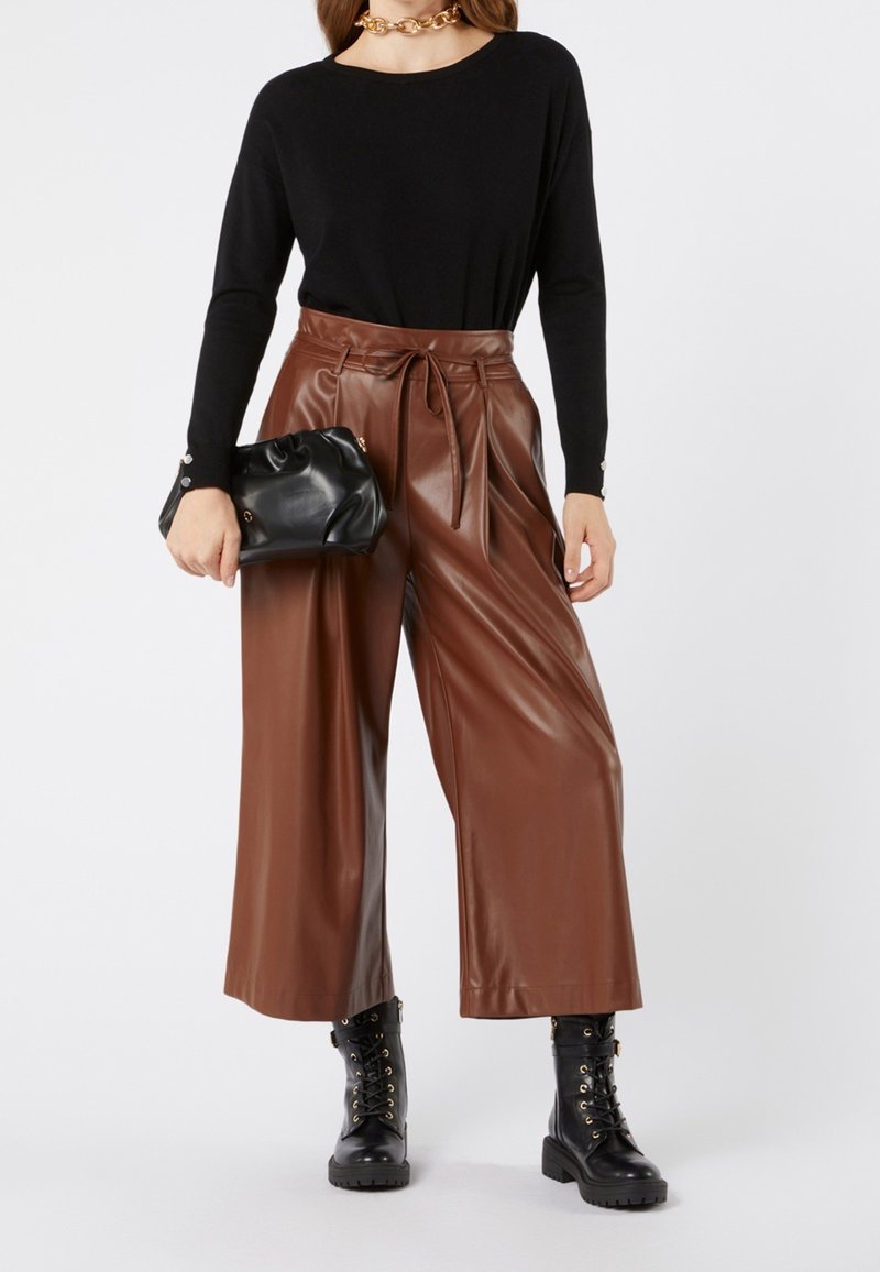 Donna con un top nero a maniche lunghe, pantaloni in pelle marroni a vita alta e a gamba larga, stivali neri con lacci, mentre tiene in mano una pochette in pelle nera.