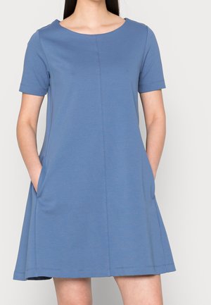 Femme portant une robe bleue à manches courtes, longueur genou, avec des poches, debout devant un fond clair uni.