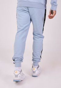 Lichtblauwe sweatpants met zwarte zijstrepen en witte tekst. Katoenen materiaal, geribbelde enkelmanchetten, gedragen met witte Nike Air sneakers.