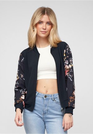 Giubbotto bomber nero con maniche a fantasia floreale, chiusura con zip e polsini a coste. Abbinato a una maglia bianca e jeans azzurri chiari.