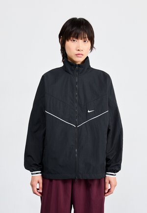 Zwarte Nike-jas met hoge kraag, rits aan de voorkant en contrasterende witte chevronstrepen. Voorzien van elastische manchetten en een lichte, gestructureerde stof.
