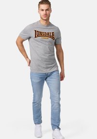 Pilkas medvilninis marškinėliai su ryškiu bordo ir aukso "LONSDALE LONDON" logo, derinami su šviesiai mėlynomis džinsomis ir baltomis kedais.