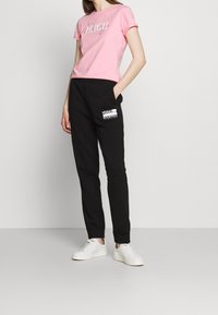 Rosa Baumwoll-T-Shirt mit "HUGO"-Logo, kombiniert mit schwarzen Jogginghosen mit Seitentasche und weißen Akzenten, abgerundet mit weißen Sneakers.