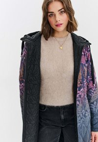 Mladá žena v béžovém huňatém svetru a květinově vzorované tmavě modré prošívané bundě, v černých džínách, stojící na bílém pozadí.