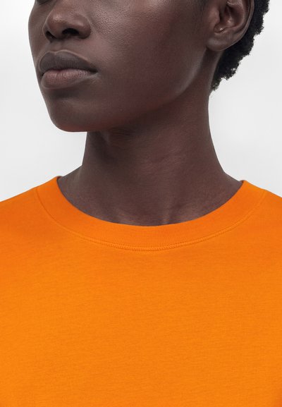 Κοντινό πλάνο του κάτω μέρους του προσώπου και του λαιμού ενός ατόμου με σκούρο δέρμα που φορά ένα φωτεινό πορτοκαλί μπλουζάκι με στρογγυλή λαιμόκοψη σε λευκό φόντο.