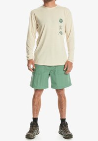 Langärmliges beiges Shirt mit grünen Grafiken, kombiniert mit grünen Shorts. Die Shorts haben einen lockeren Schnitt und eine ähnliche Stoffstruktur.
