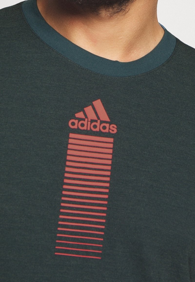 Tmavě zelené tričko s kulatým výstřihem. Na přední straně je červené logo Adidas s vertikálním pruhovaným designem. Měkká textilie.