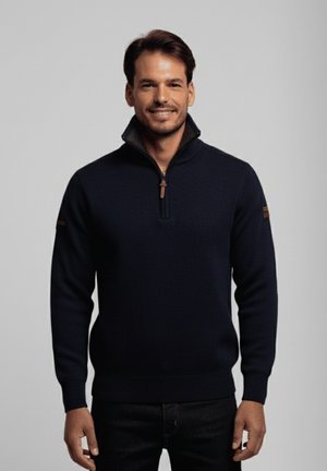 Hombre sonriente con cabello oscuro que lleva un suéter azul marino con cremallera y jeans oscuros, de pie contra un fondo gris claro liso.