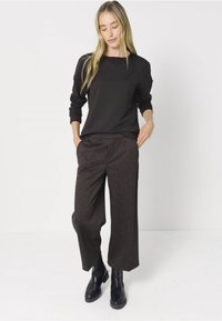 Nainen, joka on pukeutunut mustaan pitkähihaiseen paitaan, tummiin raidallisiin leveälahkeisiin housuihin ja mustiin nilkkabootseihin, seisoo kädet taskuissa.