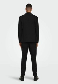 Czarny garnitur z dopasowaną marynarką z wycięciem, prostymi spodniami oraz błyszczącymi czarnymi butami, wszystko pokazane z tyłu.